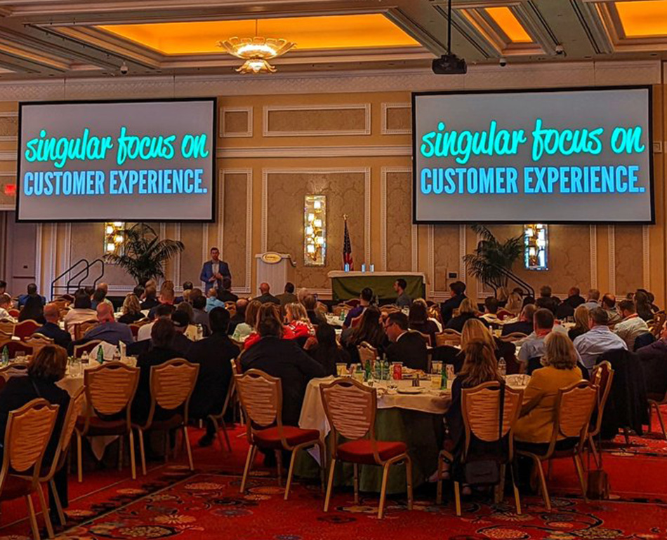 Large ballroom full of people with two large screens displaying the text, Singular Focus on Customer Experience